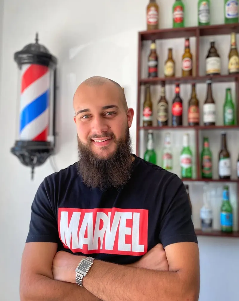 Pedro Caramujo, barbeiro, de braços cruzados na barbearia, com o poste de barbeiro e a parede de garrafas atrás